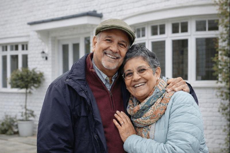 Smiling older couple, leaning on each other and standing outside their home