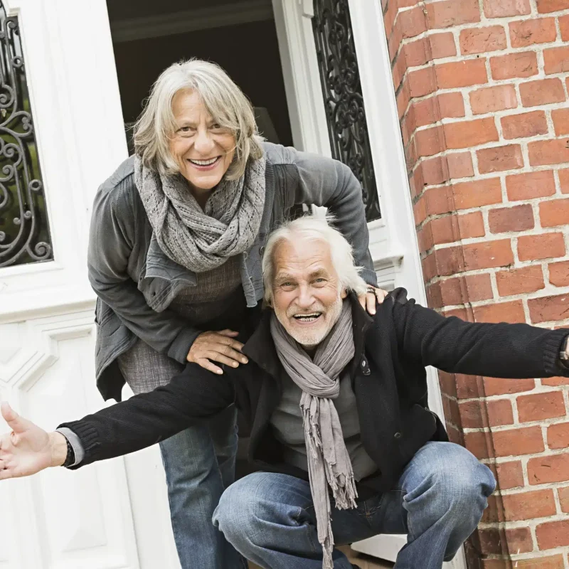 senior-couple-by-front-door-man-crouching-with-op-2024-06-04-17-16-37-utc